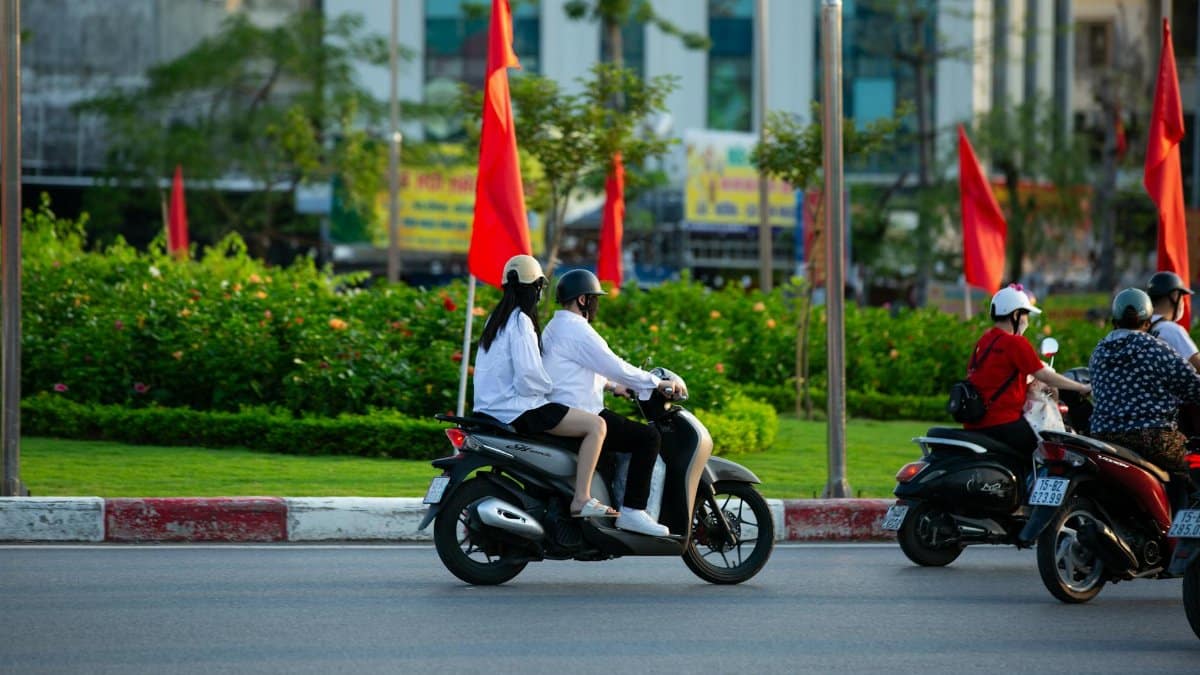 A group of people riding scooters on an urban road lined with red flags, indicating a vibrant city scene.