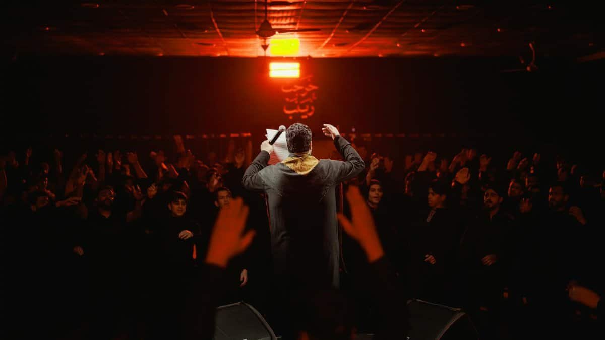 Back view of a performer engaging a lively crowd under dramatic lighting in a dark, indoor setting.