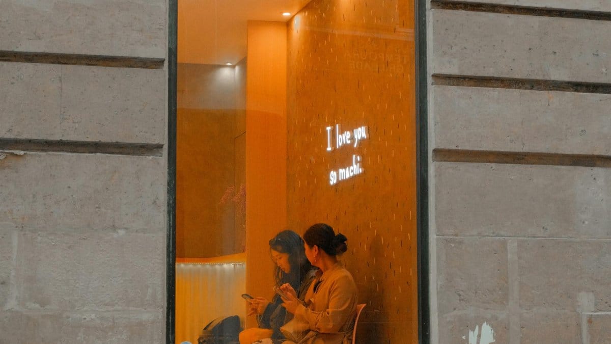 Two women sit in a cozy cafe, viewed through a window with a neon 'I love you so much' sign.