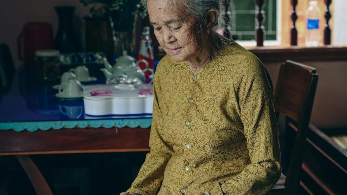 An elderly Asian woman in a green gown sitting indoors, conveying wisdom.