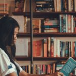 A young woman reads a book in a comfortable library with shelves full of books, offering a serene setting for relaxation.