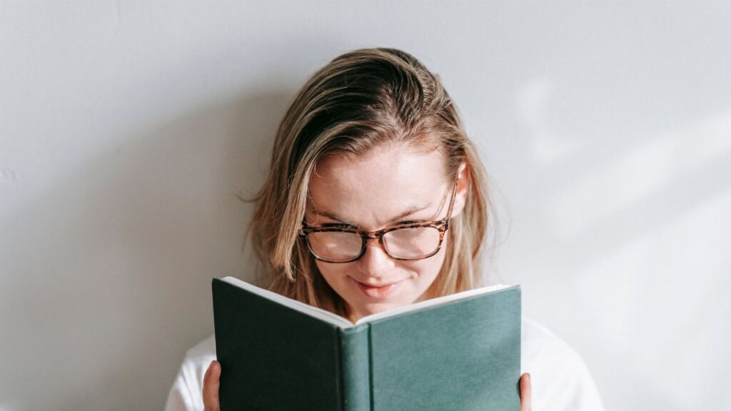 Interested young woman in glasses reading textbook while studying in sunshine on light background