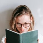 Interested young woman in glasses reading textbook while studying in sunshine on light background