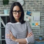 Portrait of a woman with glasses standing confidently in a modern office setting.