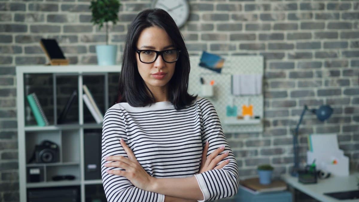 Portrait of a woman with glasses standing confidently in a modern office setting.