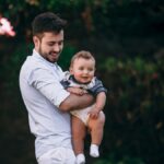 A joyful father holds his baby while standing in a lush garden, capturing a heartwarming outdoor moment.