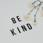 Minimalist image of 'Be Kind' text with white flowers on a light background, promoting kindness.