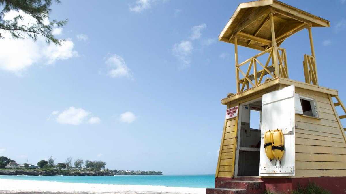 A wooden lifeguard tower stands on a sunny Barbados beach with clear blue skies and ocean.