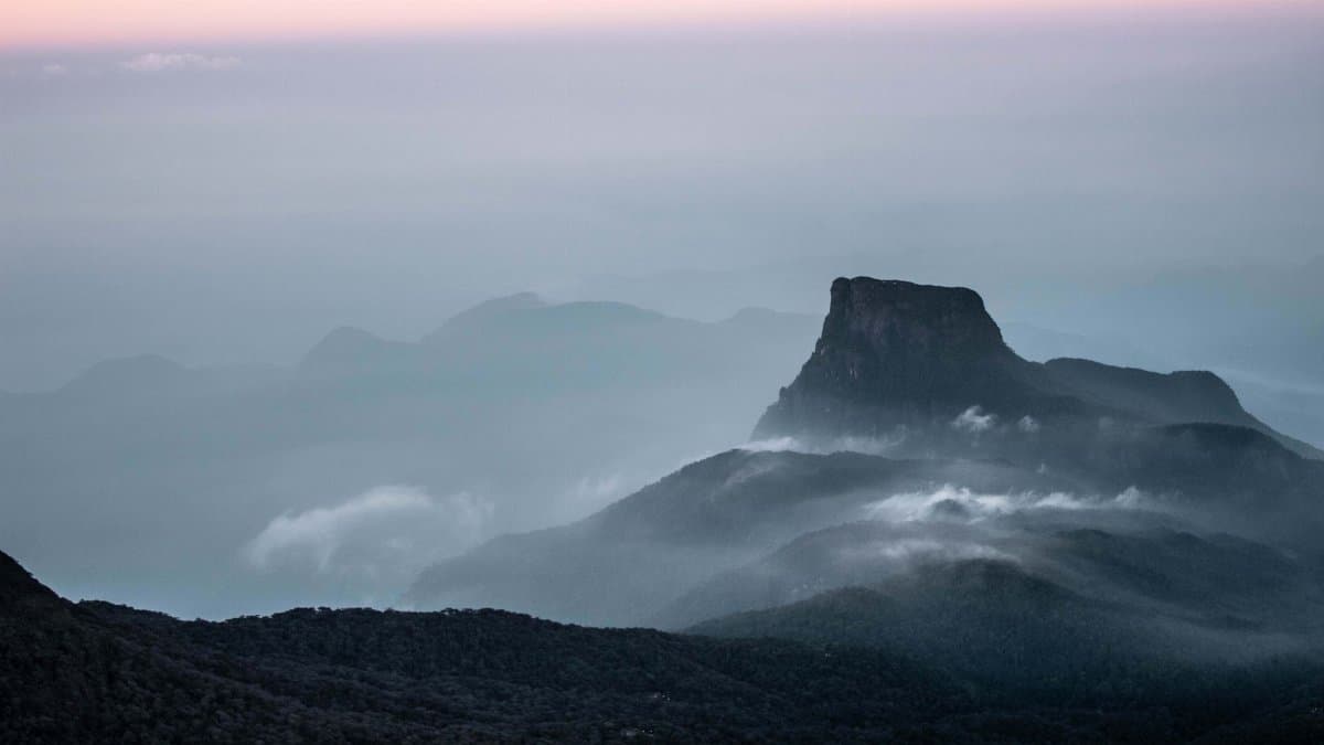 Serene view of Adam's Peak at dawn, shrouded in mist and gentle clouds, in Sri Lanka's lush landscape.