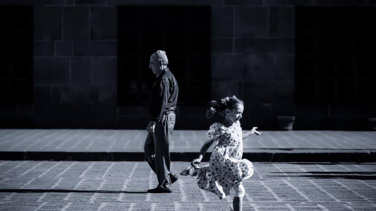 A young girl in a sundress running on a pavement with an elderly man walking nearby in an urban setting.