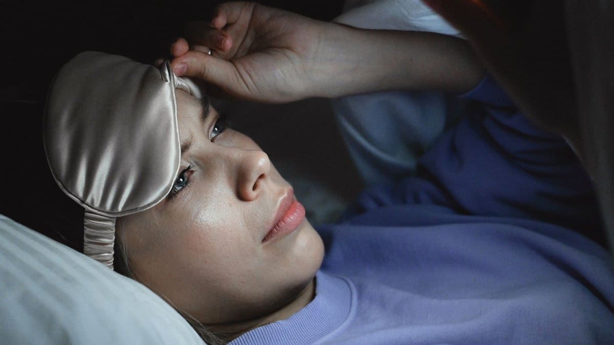 A woman in bed peeking under a sleep mask to use her smartphone at night, creating a relaxing bedtime scene.