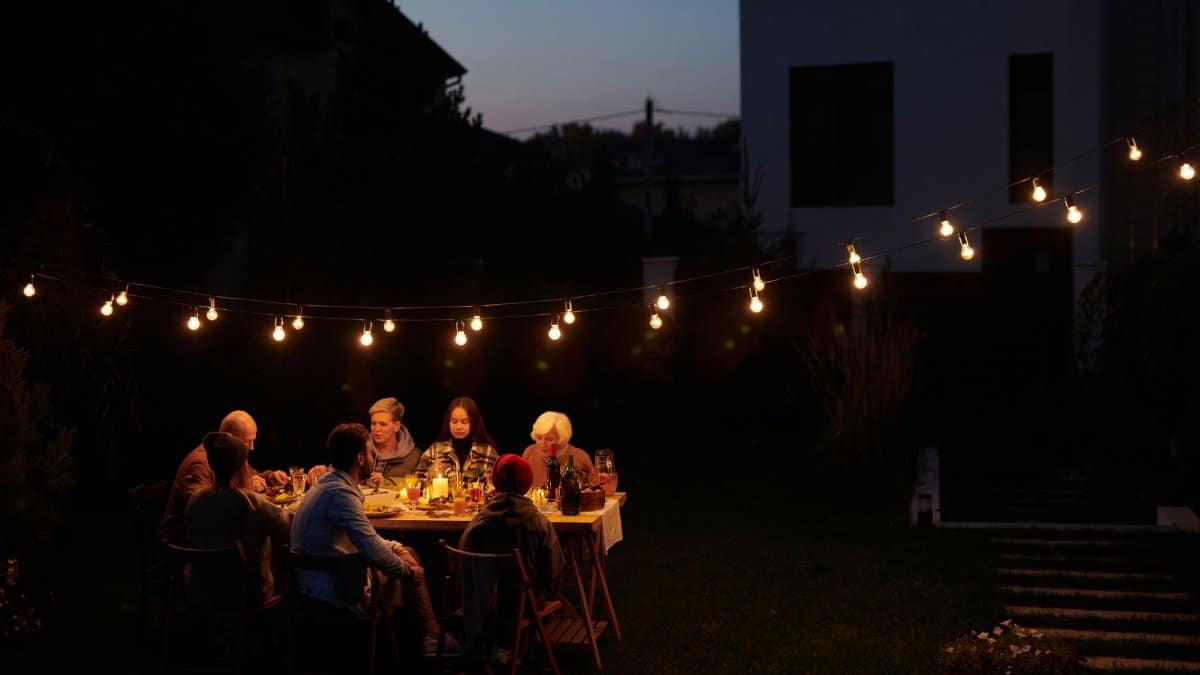 A family enjoys an evening meal outdoors under decorative string lights in a garden setting.