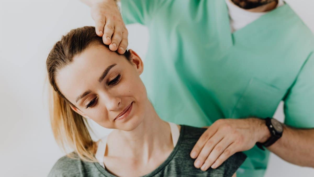 Serious doctor in uniform massaging smiling female patients neck and stretching stiff neck muscles against gray background