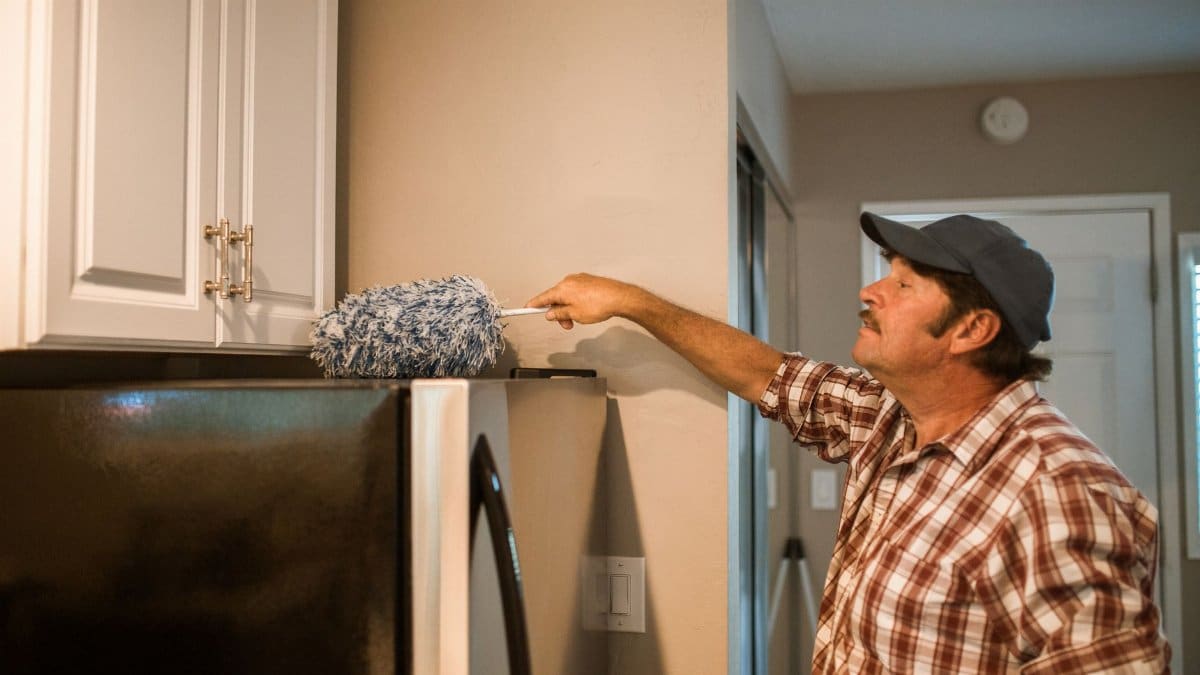 Middle-aged man using a duster to clean kitchen cabinets at home.