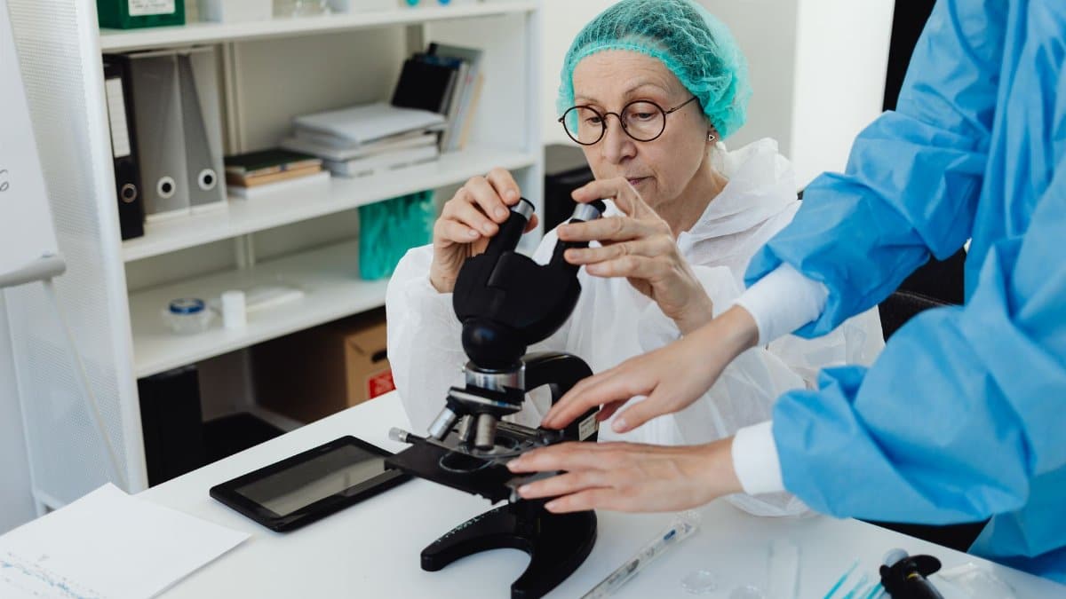 Senior researcher working with a microscope in a laboratory setting, assisted by a colleague.