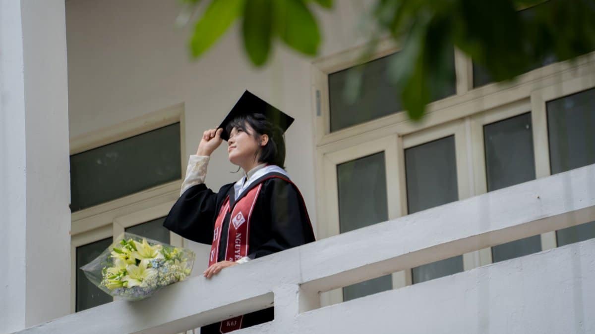 Graduate stands proudly with flowers and cap, celebrating achievement outdoors.