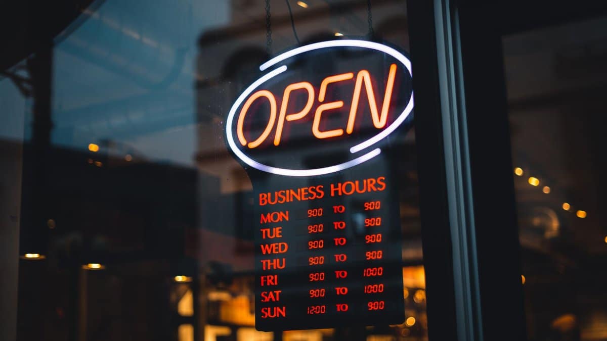 A glowing neon 'OPEN' sign with detailed business hours in a storefront window at night.