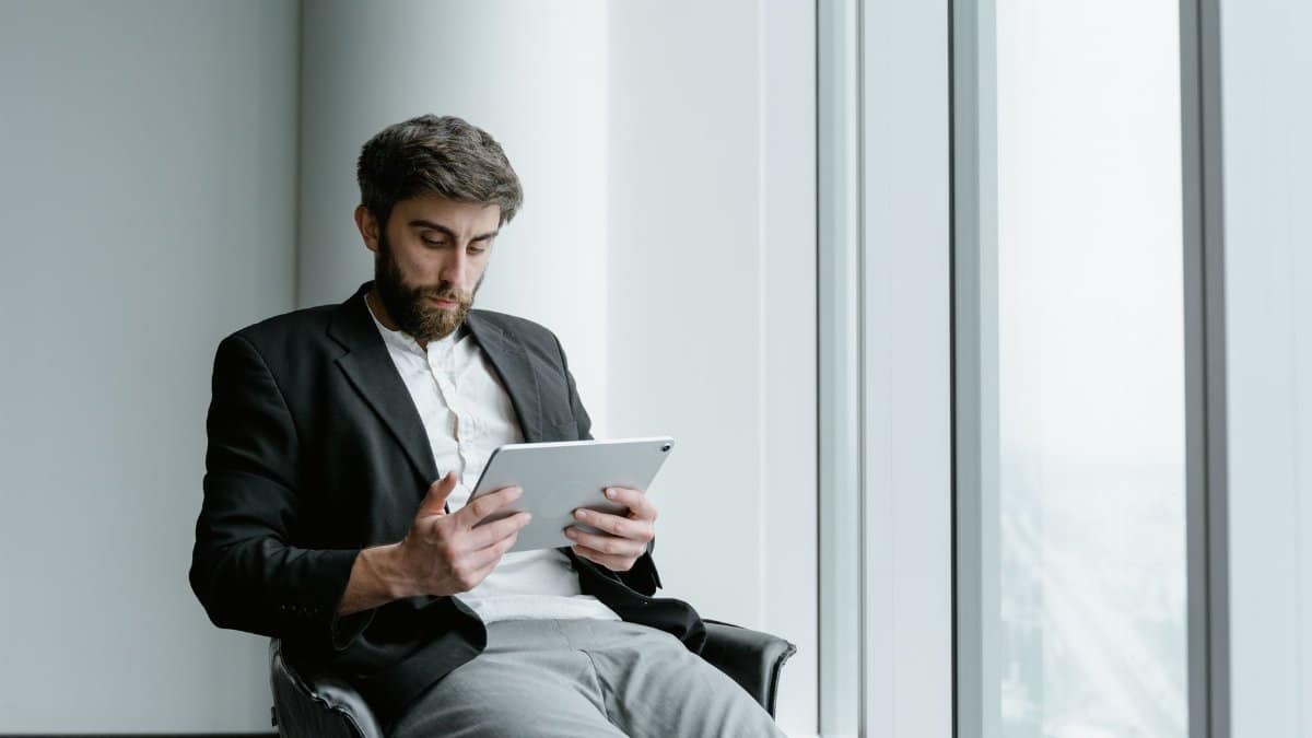 A man in formal attire working on a tablet while seated by a large office window.