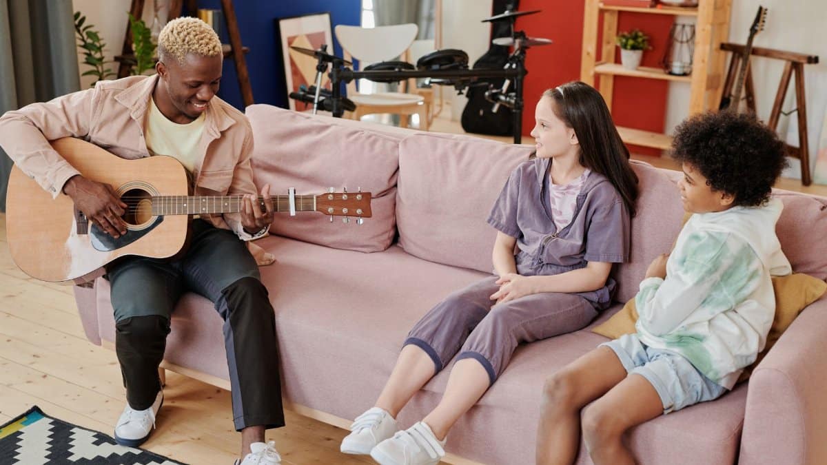Man teaching guitar to children on a sofa in a cozy living room.
