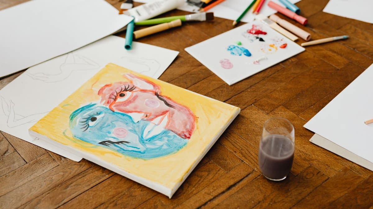 Art supplies scattered on a wooden floor, featuring vibrant paintings and tools for creative projects.