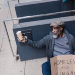 A homeless man sitting on a sidewalk holding a cup and sign in an urban area.