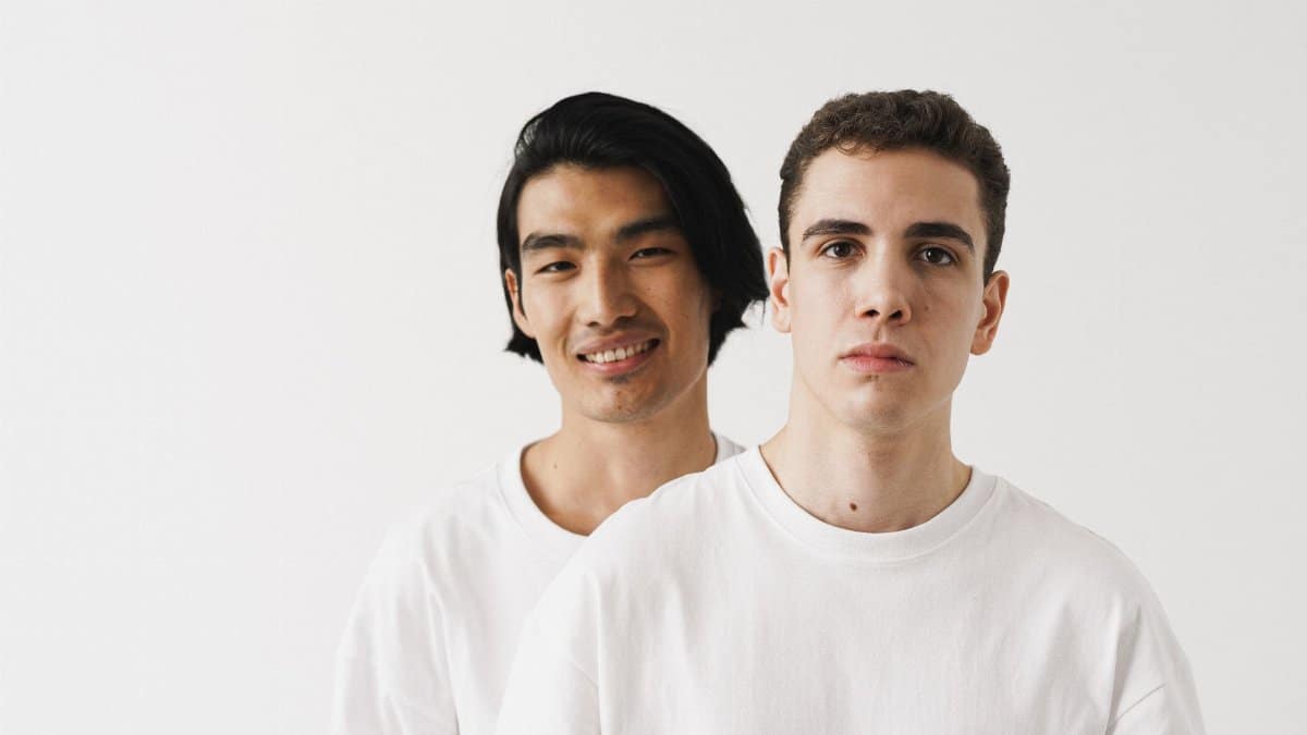 A close-up portrait of two men wearing white shirts against a neutral background.