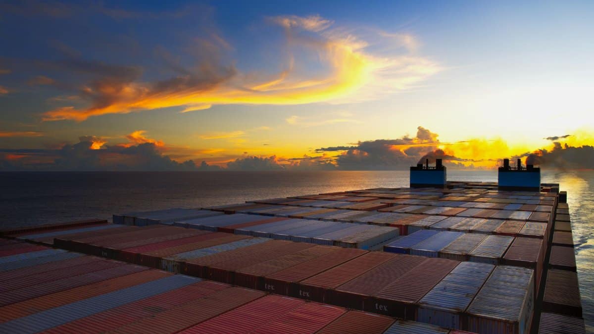 Aerial view of colorful shipping containers on a vessel against a stunning sunset sky over the ocean.