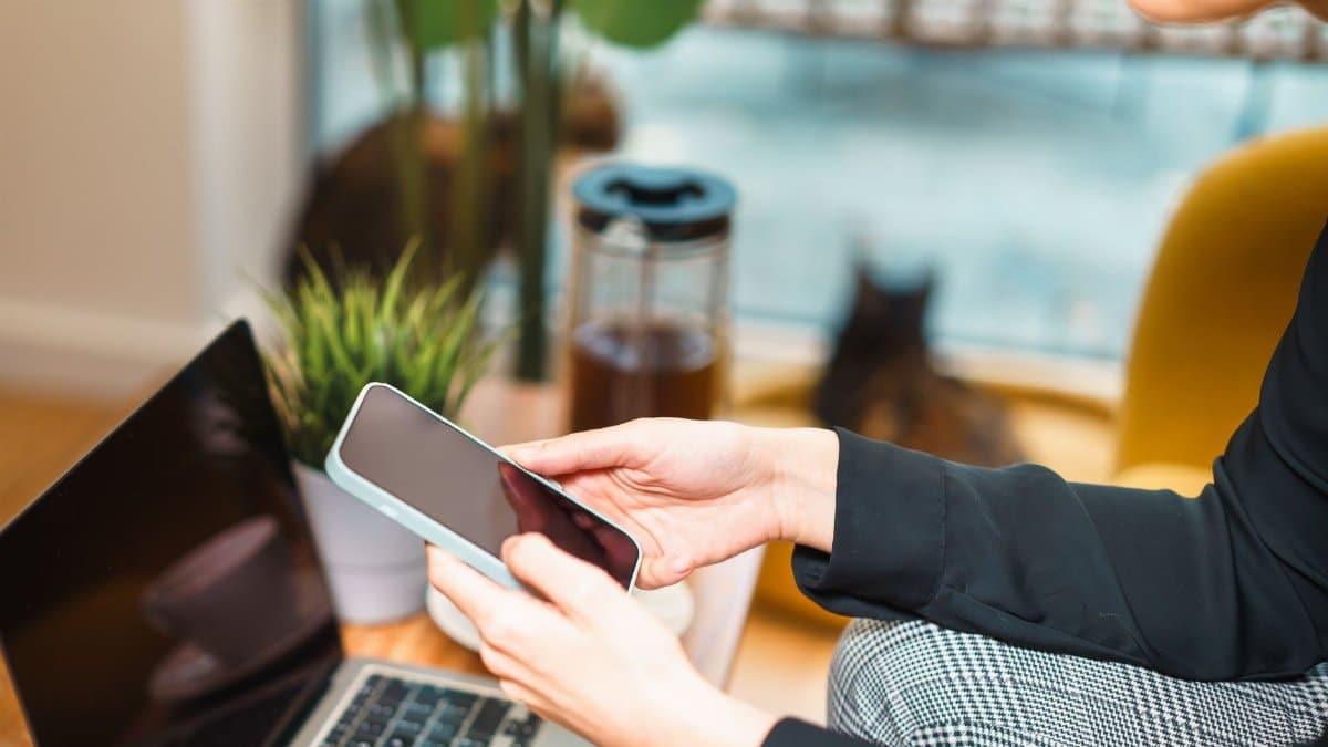 Woman working remotely from home, using a smartphone and laptop at a cozy desk with a plant and cat in background.