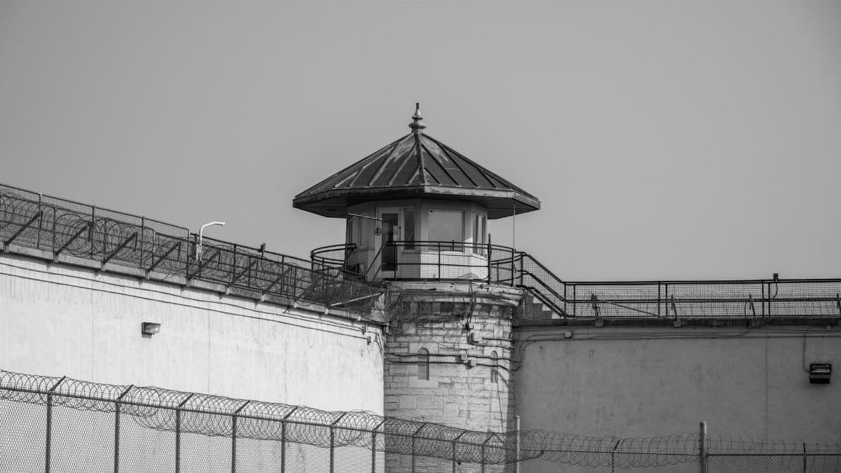 Black and white image of a prison watchtower with barbed wire fencing and security measures under a clear sky.