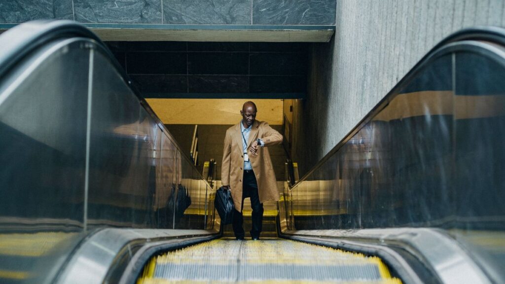 High angle full length pensive African American businessman in formal clothes with laptop case riding escalator and checking time on wristwatch while leaving metro station