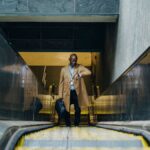 High angle full length pensive African American businessman in formal clothes with laptop case riding escalator and checking time on wristwatch while leaving metro station