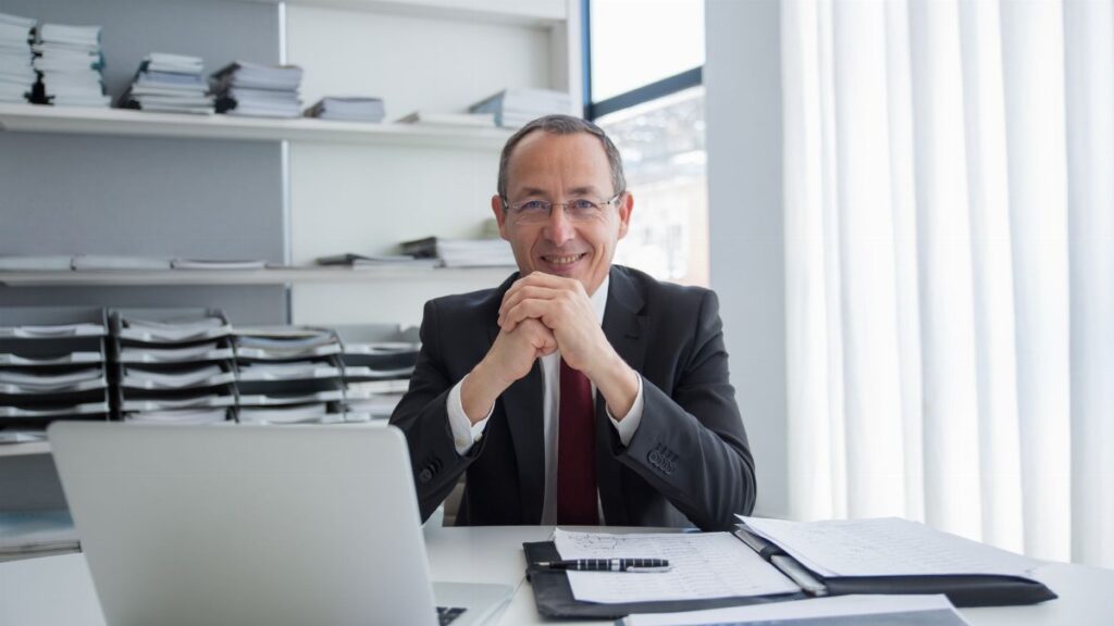 Smiling businessman in a suit with laptop and documents, exuding confidence.