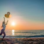 A woman joyfully running with balloons along a beach at sunset, capturing a moment of happiness.