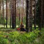 A woman sits alone in a lush, dense forest, surrounded by tall trees in daylight.