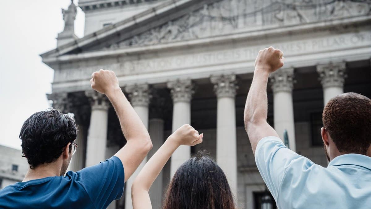Three people raising fists in protest outside a government building with Greek-style architecture.