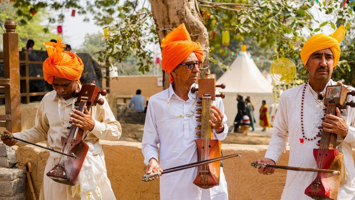 Three Rajasthani musicians play traditional instruments outdoors, wearing vibrant turbans.