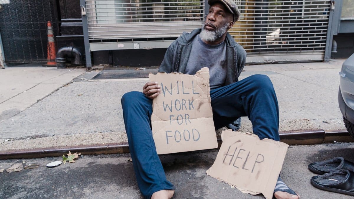 Homeless man sitting on sidewalk holding signs for help and work on a city street.