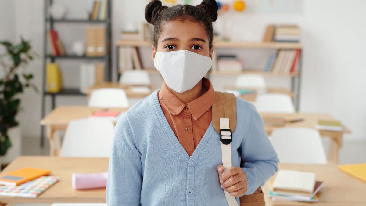 Girl with backpack and face mask standing in a classroom, symbolizing back-to-school safety.