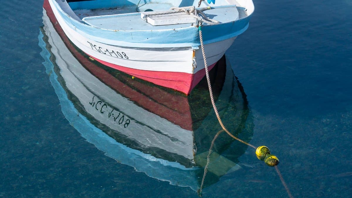 A small fishing boat with vibrant colors reflected on calm, clear water tied with rope.