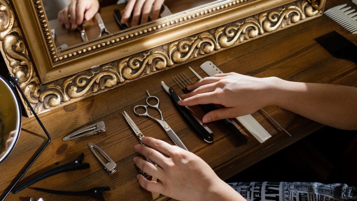 Hands arranging hair styling tools on a wooden table near a decorative mirror.