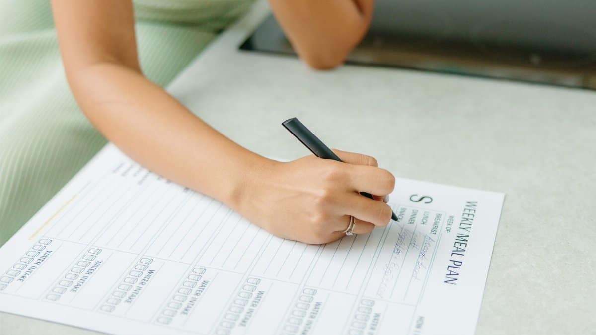 Close-up of a woman writing a weekly meal plan at home, promoting a healthy lifestyle.