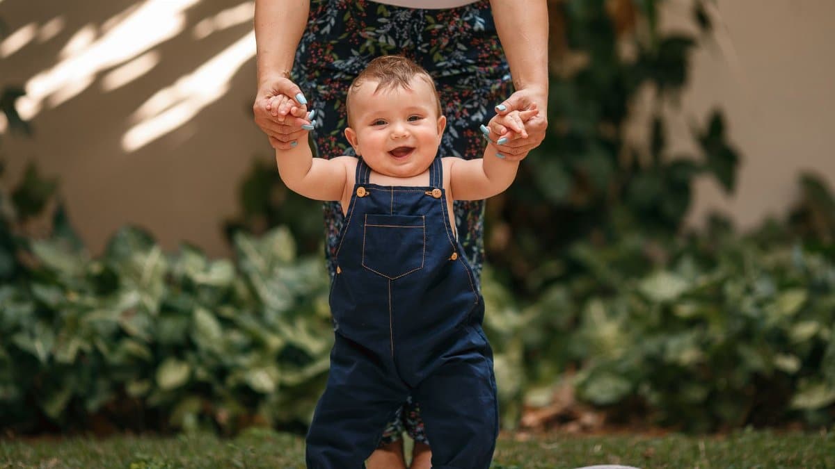 Cheerful baby in overalls being supported while taking first steps outdoors.