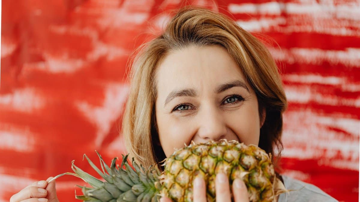 Cheerful woman playfully hides behind a pineapple against a vibrant red background.