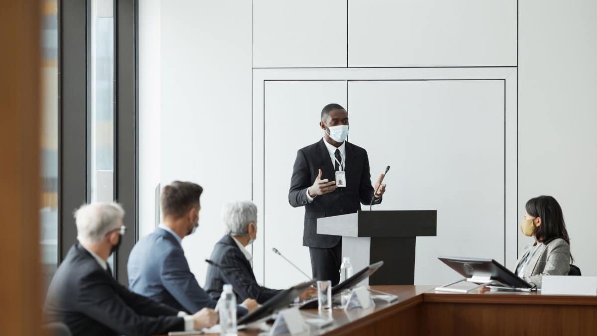 Business professionals in masks hold a meeting indoors, highlighting new normal practices during pandemic times.