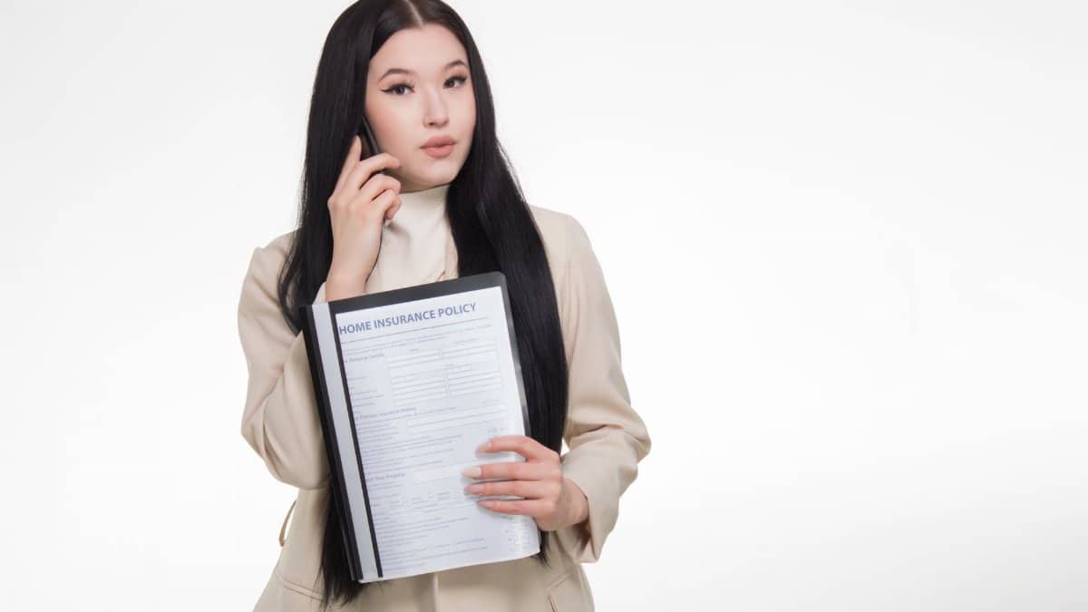 Asian woman in business attire holding home insurance policy while on a phone call.