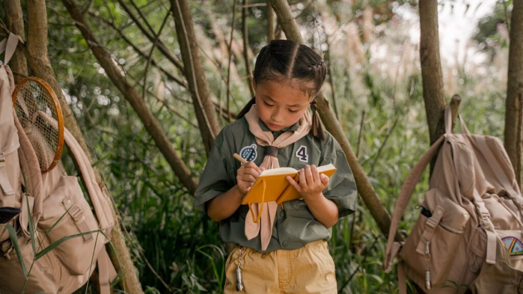 A young girl scout takes notes during a summer camp in the woods.