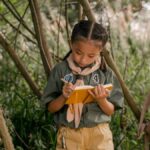 A young girl scout takes notes during a summer camp in the woods.