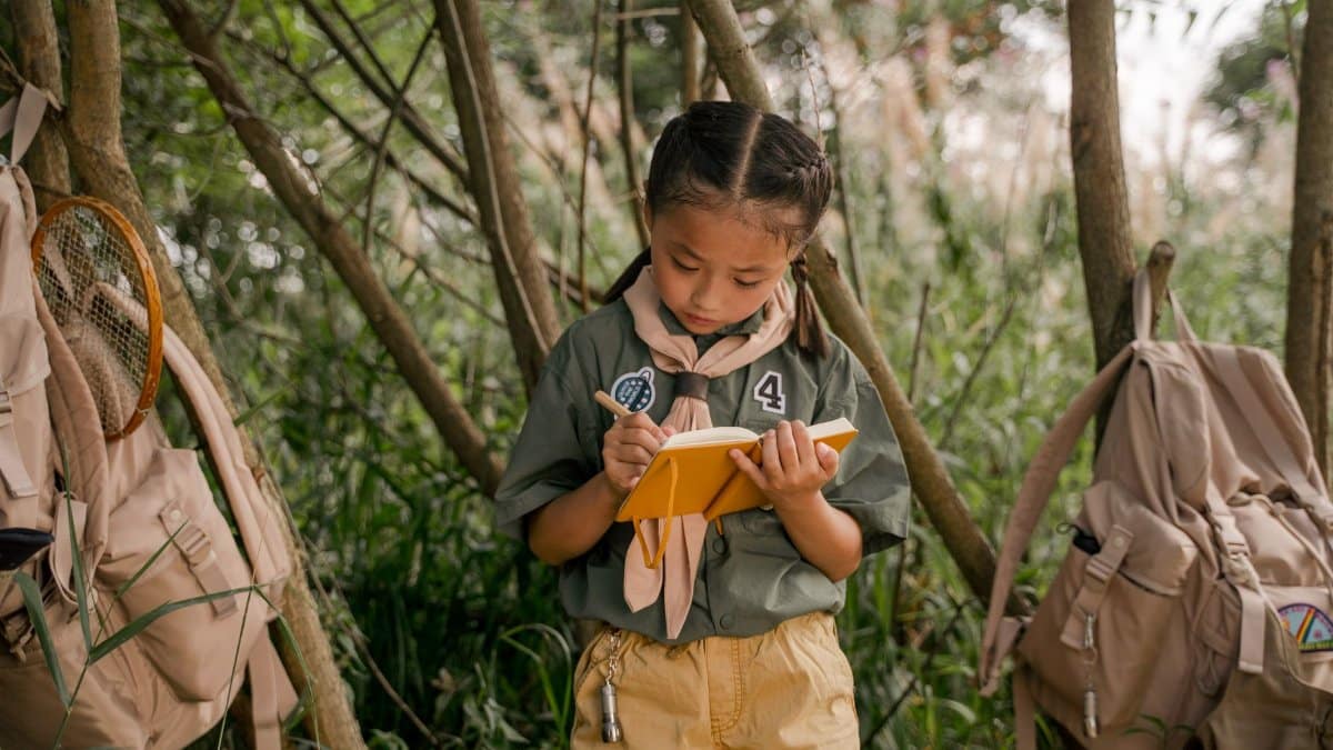 A young girl scout takes notes during a summer camp in the woods.