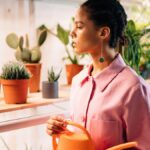 A woman in a pink shirt waters indoor potted plants in a modern studio.