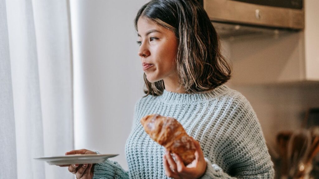 Attentive Asian female standing near curtains with delicious croissant in kitchen and looking away